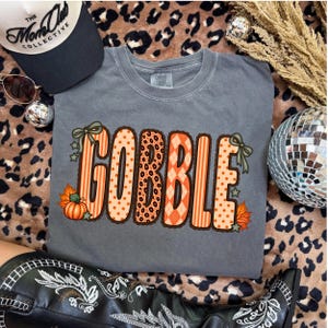 K&ouml;nnte beinhalten: Ein anthrazitfarbenes T-Shirt mit dem Wort GOBBLE in gro&szlig;en, orangefarbenen Buchstaben mit verschiedenen Mustern wie Streifen, Punkten, Leopardenmuster und Rauten. Das Design ist mit kleinen gr&uuml;nen Schleifen, Sternen und orangefarbenen K&uuml;rbissen verziert.