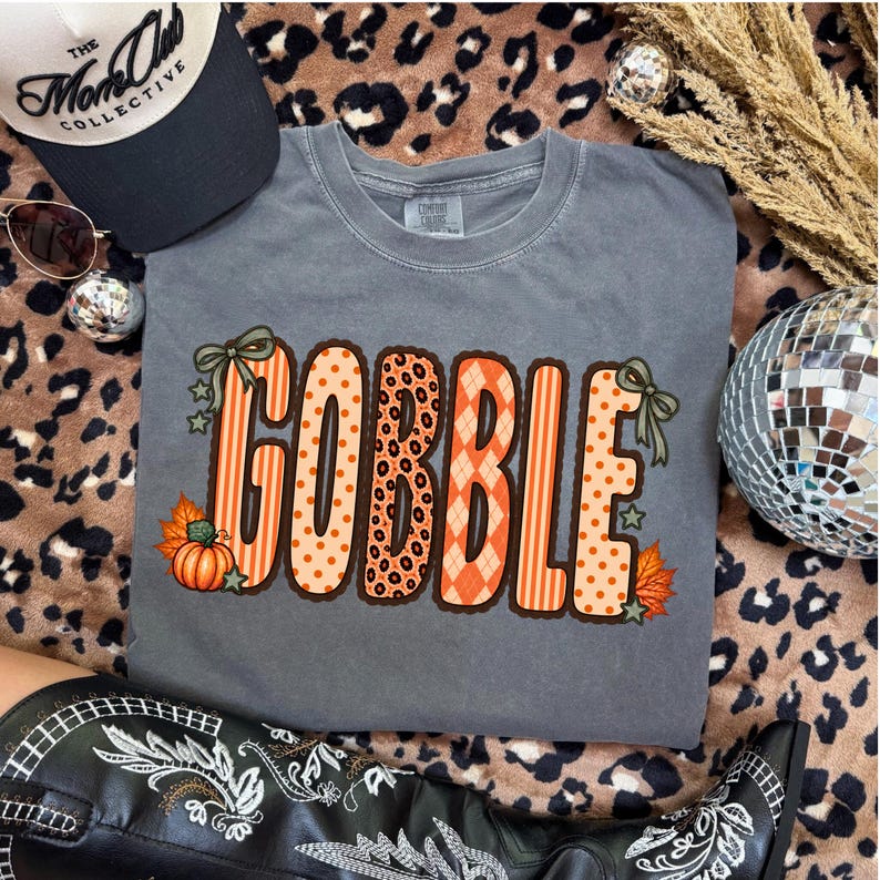 K&ouml;nnte beinhalten: Ein anthrazitfarbenes T-Shirt mit dem Wort GOBBLE in gro&szlig;en, orangefarbenen Buchstaben mit verschiedenen Mustern wie Streifen, Punkten, Leopardenmuster und Rauten. Das Design ist mit kleinen gr&uuml;nen Schleifen, Sternen und orangefarbenen K&uuml;rbissen verziert.