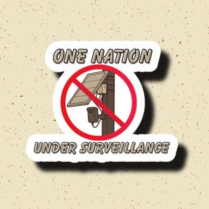 May include: A sticker featuring a graphic of a solar panel and a security camera mounted on a pole, crossed out by a red circle. The text reads ONE NATION above and UNDER SURVEILLANCE below in a bold, light-coloured font with dark outlines.