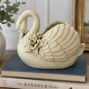Pode incluir: Um vaso de cerâmica cor de creme em forma de cisne, com uma rosa esculpida no peito e texturas de penas detalhadas nas asas. O cisne possui um pescoço curvo e uma abertura superior, apresentando um acabamento brilhante.