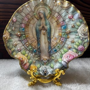 May include: A decorative scallop shell featuring a central illustration of a figure in a blue and white robe with hands clasped in prayer. The shell is adorned with colorful rhinestones and floral motifs, set on a gold-toned metal display stand.