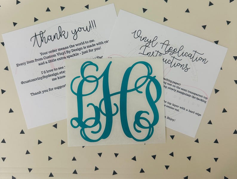 May include: A teal monogram vinyl decal with the letters "JH" in a decorative script. The decal is centered on a white square. Two white cards with handwritten text, including "Thank you!!!" and "Vinyl Application Instructions", are in the background.