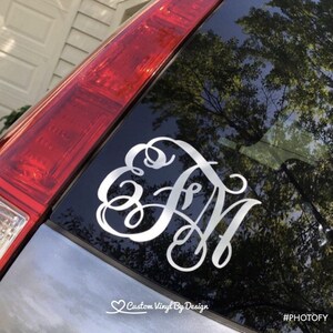 May include: A white, cursive monogram decal, featuring the letters E, F, and N, is affixed to a car window. The decal is a custom vinyl design. The image also includes the text "Custom Vinyl By Design" and the hashtag "#PHOTOFY".