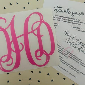 May include: A hot pink monogram decal with the letters DH in a cursive font. Accompanying the decal are two printed cards with instructions and a thank you message. The background is a white surface with black triangles.