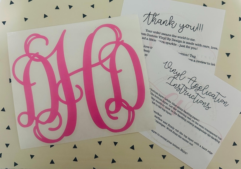 May include: A hot pink monogram decal with the letters DH in a cursive font. Accompanying the decal are two printed cards with instructions and a thank you message. The background is a white surface with black triangles.
