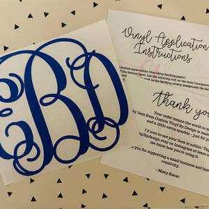 May include: A blue monogram vinyl decal with the letters EBD, displayed on a white background. Accompanying the decal are two cards with handwritten text, including application instructions and a thank you note. The background has a pattern of black triangles.