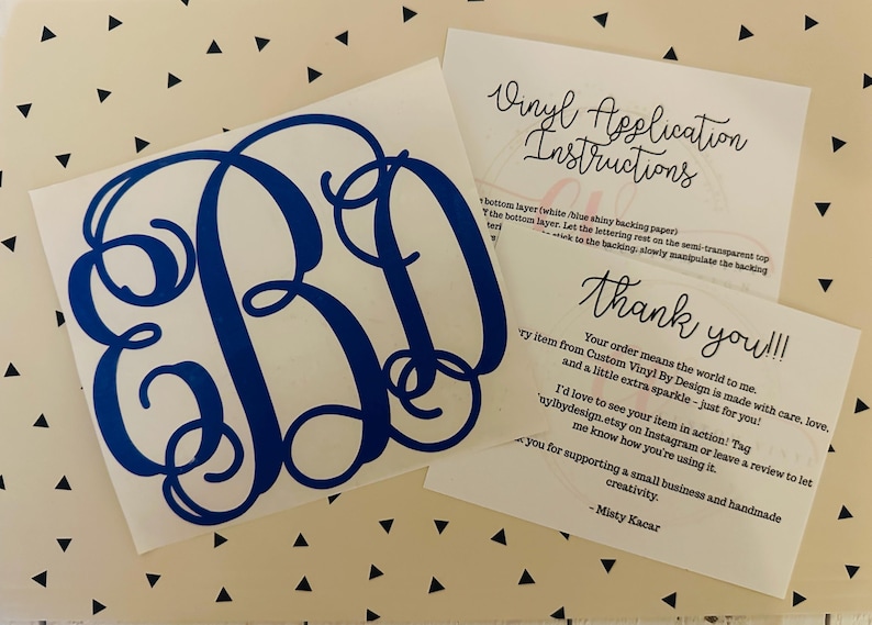 May include: A blue monogram vinyl decal with the letters EBD, displayed on a white background. Accompanying the decal are two cards with handwritten text, including application instructions and a thank you note. The background has a pattern of black triangles.