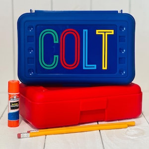 May include: A blue rectangular plastic pencil box with the name COLT printed in green, red, blue, and yellow letters. Below it sits a red plastic pencil box, an orange glue stick, and two yellow wooden pencils with pink erasers.