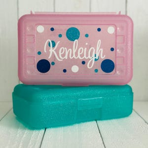 May include: A rectangular pink glittery plastic pencil case with a teal and white polka dot pattern and the name Kenleigh written in white script font, stacked on top of a solid teal glittery plastic pencil case with a rectangular latch.