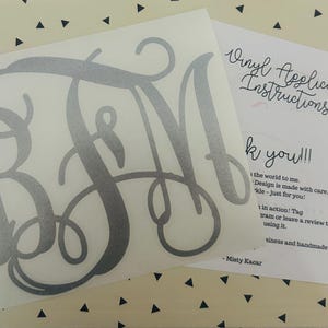 May include: A silver monogram decal with the letters BFM in a cursive font. The decal is on a white background, with a sheet of paper next to it that says "Vinyl Application Instructions" and "Thank you!!!".