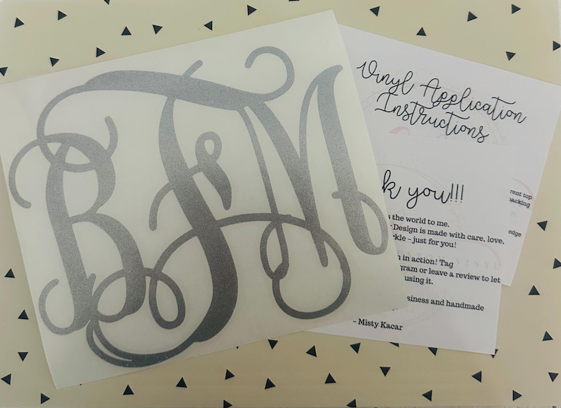 May include: A silver monogram decal with the letters BFM in a cursive font. The decal is on a white background, with a sheet of paper next to it that says "Vinyl Application Instructions" and "Thank you!!!".