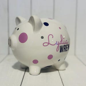 May include: A white ceramic piggy bank with a glossy finish. It features purple and navy polka dots and the name "Lydia Wren" in a cursive font. The piggy bank has four small feet and a slot on top.