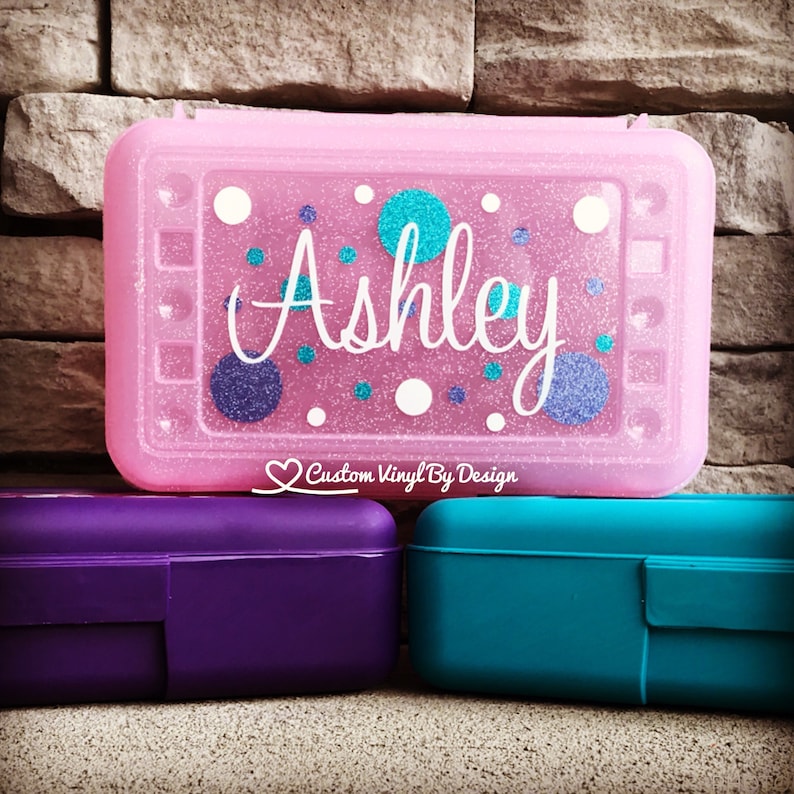 May include: A rectangular pink glittery pencil case with the name Ashley in white script, decorated with teal and purple glittery polka dots. Below it are two solid-coloured pencil cases, one purple and one teal, with a small logo reading Custom Vinyl By Design.