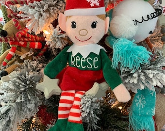 Puede incluir: Un muñeco de elfo de peluche con un gorro rojo con un copo de nieve blanco, una camisa verde con el nombre "Reese" y una falda roja. El elfo tiene piernas a rayas rojas y blancas y pies verdes con el año "2019". El muñeco es parte de una decoración navideña.