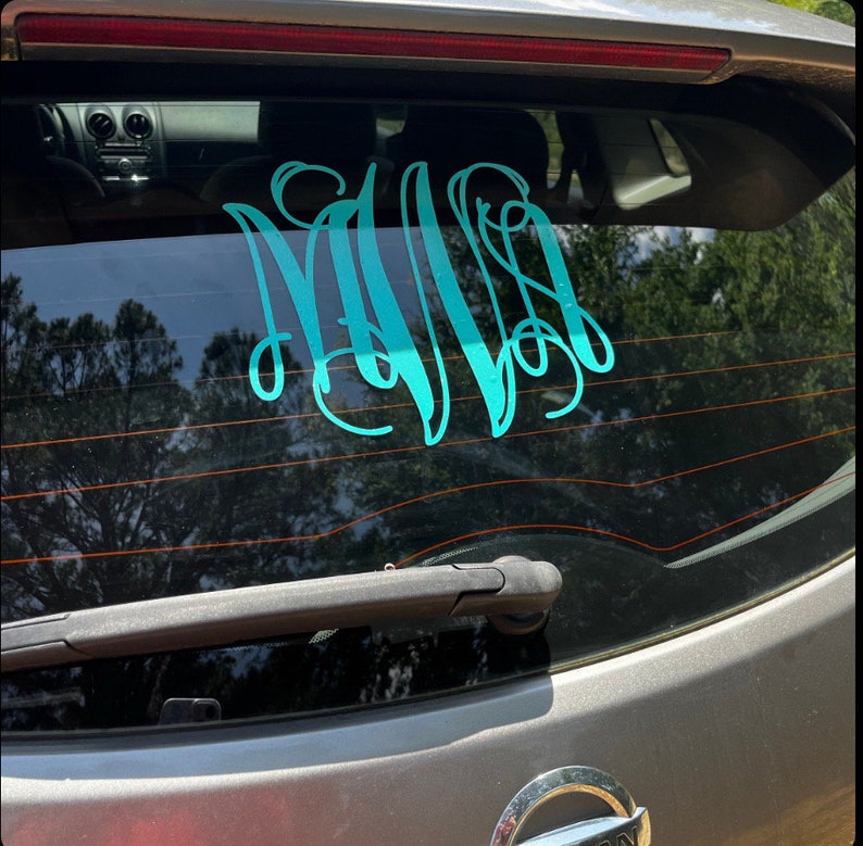 May include: A teal monogram decal is affixed to the rear window of a gray vehicle. The decal features three intertwined cursive letters, creating a personalized and decorative element. The car's wiper blade is visible.