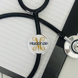 May include: A white stethoscope charm with a gold initial "M" and the name "Mackenzie" in navy blue lettering.