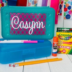 May include: A turquoise plastic pencil case with a pink and white patterned design and the name "Casyun" on the lid. The case is open and contains two markers, two pencils, and a glue stick. A box of 24 Crayola crayons is also visible.