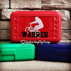 May include: Red lunchbox with a white silhouette of a person riding a BMX bike and the name "WARREN" in black. The lunchbox is rectangular with rounded corners. Two other lunchboxes, green and blue, are in the foreground.
