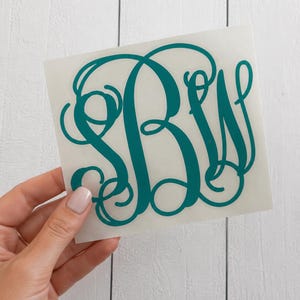 May include: Teal monogram sticker featuring the letters B, W, and a stylized initial. The sticker is held against a white wooden background. The letters are in a cursive font.