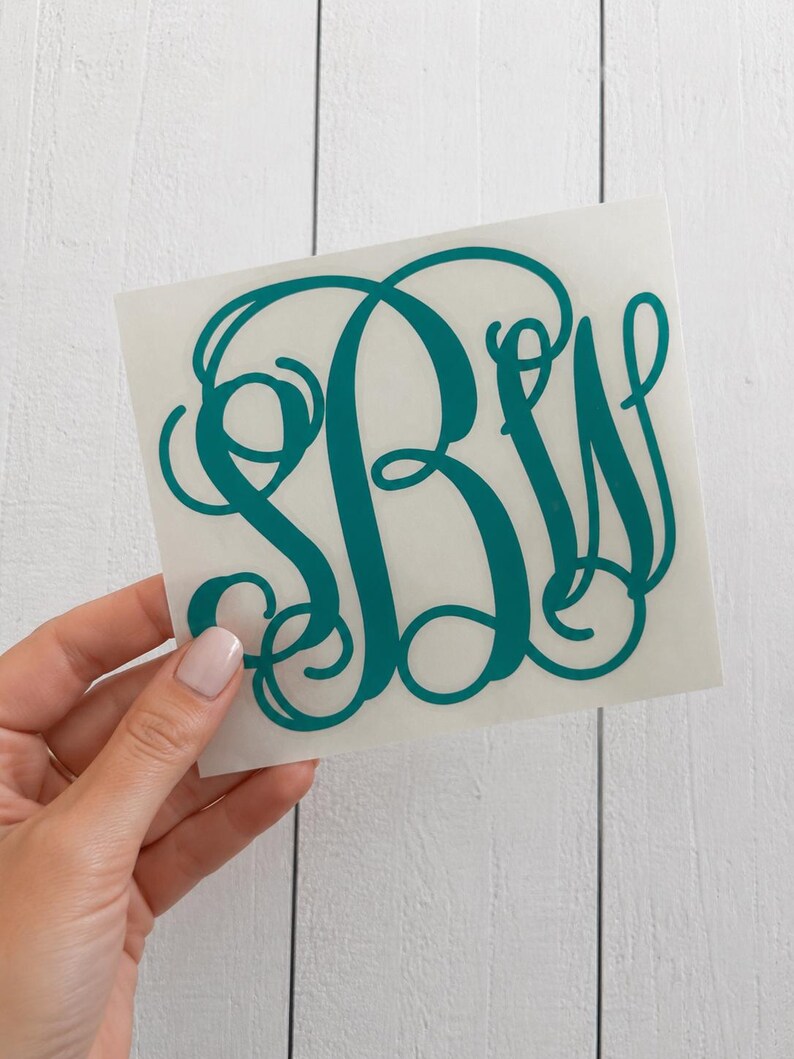 May include: Teal monogram sticker featuring the letters B, W, and a stylized initial. The sticker is held against a white wooden background. The letters are in a cursive font.