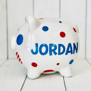 May include: White ceramic piggy bank with red and blue polka dots. The name "JORDAN" is written in blue letters on the side.