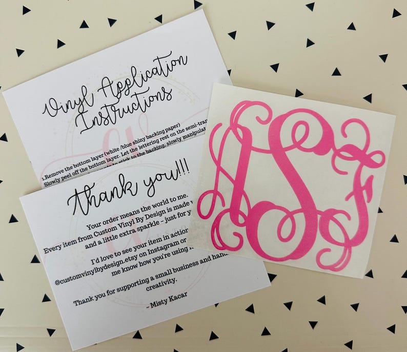 May include: A hot pink monogram vinyl decal with a cursive font, next to two white cards with black text. One card reads "Vinyl Application Instructions" and the other says "Thank you!!!". The background is a cream color with black triangles.