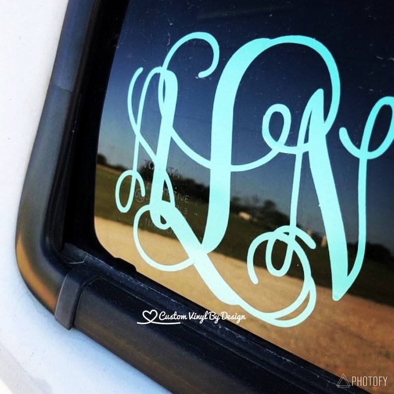 May include: A teal monogram decal, featuring three intertwined letters, is affixed to a car window. The decal is in a cursive font, with a heart and the text "Custom Vinyl By Design" at the bottom. The background shows a glimpse of a field.