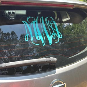 May include: A teal monogram decal is affixed to the rear window of a gray vehicle. The decal features three intertwined cursive letters. The car's wiper blade is visible at the bottom, and trees are reflected in the window.