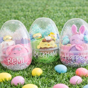 May include: Three clear plastic Easter egg containers filled with gifts, each with a name on the front: Remi, Beckham, and Harper. The eggs sit on green grass with colorful Easter eggs scattered around. Each egg contains a plush toy and decorative accents.