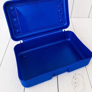 May include: A rectangular, open plastic storage box in a vibrant royal blue colour. The lid is attached with a hinge and features a recessed rectangular panel surrounded by small circular and square moulded indentations along the inner border.