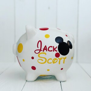 May include: White ceramic piggy bank with red and yellow polka dots. The piggy bank has a black Mickey Mouse head silhouette and the name "Jack Scott" written in red and yellow.