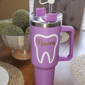 May include: A purple stainless steel tumbler with a white tooth graphic and the name "Raven" in gold lettering. The tumbler has a lid with a straw hole and a handle.
