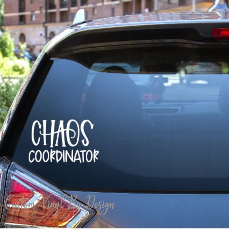 May include: White vinyl decal on a car window that reads "CHAOS COORDINATOR" in a whimsical font. The decal is applied to the rear window of a vehicle, with a blurred background of buildings and sky.
