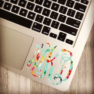May include: A floral monogram sticker in shades of pink, green, and blue, placed on a silver laptop.