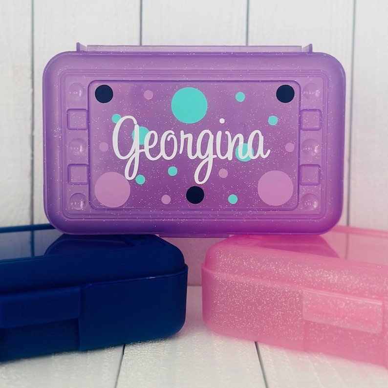 May include: A rectangular purple glittery plastic pencil case featuring the name Georgina in white script lettering. The lid is decorated with various sized circles in shades of teal, dark blue, and light purple against the translucent purple base.