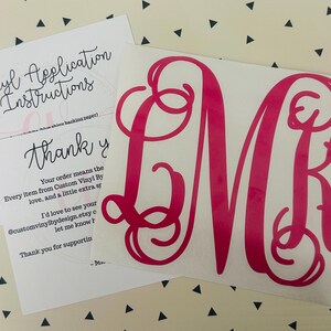 May include: A hot pink monogram decal with the letters LMK, accompanied by vinyl application instructions and a thank you note. The decal is on a white backing, and the text is in a cursive font. The background is a cream color with black triangles.