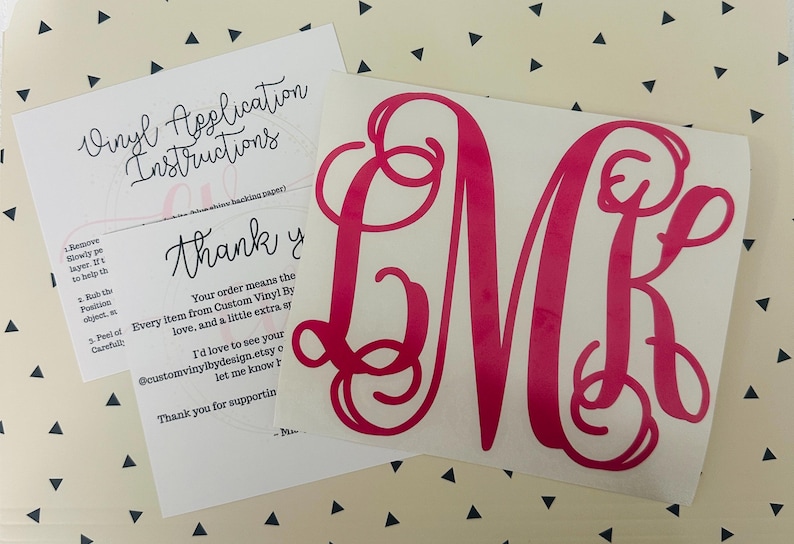 May include: A hot pink monogram decal with the letters LMK, accompanied by vinyl application instructions and a thank you note. The decal is on a white backing, and the text is in a cursive font. The background is a cream color with black triangles.