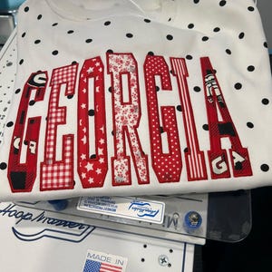 May include: A white sweatshirt featuring black polka dots and the word GEORGIA appliqued in red-bordered letters. Each letter displays a unique red and white pattern, including plaid, stars, floral, and stripes, with a bulldog mascot graphic.
