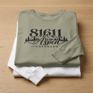 May include: A sage green long-sleeve shirt layered over a white long-sleeve shirt. The sage shirt features a black graphic with the text 81611, a mountain range silhouette, a forest of pine trees, and the word Aspen Colorado in script font.