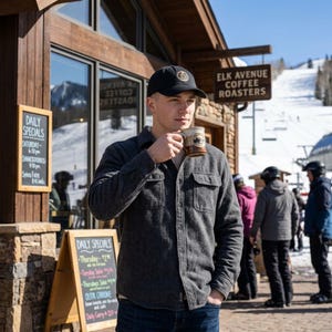 May include: A black baseball cap with a circular logo, a dark grey long-sleeve button-down shirt with two chest pockets, and a two-toned brown and cream ceramic mug with a circular logo held in front of a coffee shop with snowy mountains.