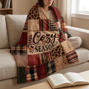 May include: A patchwork-style throw blanket featuring various textures and patterns including leopard print, cable knit, plaid, and houndstooth in shades of brown, red, and cream. The center displays the text Cozy SEASON with a small heart icon.