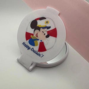 May include: A white circular folding compact mirror featuring a graphic of a cartoon mouse wearing a captain's hat, set against a red and white life preserver design. The text Happy Cruisin'! is printed in blue script on the front cover.