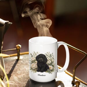 May include: A white ceramic mug featuring a graphic illustration of a black puppy sitting amidst a cluster of white flowers and green foliage. The name Shadow is printed in black text below the puppy. Steam rises from the top of the mug.