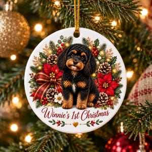 May include: A round white ceramic Christmas ornament featuring a black and tan puppy sitting in the center. The puppy is surrounded by a festive wreath of pine branches, red poinsettias, pinecones, and berries. The text Winnie's 1st Christmas is printed in red script at the bottom with a small gold heart.