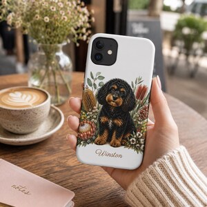 May include: A white smartphone case featuring a detailed illustration of a black and tan puppy sitting among various colorful flowers and greenery. The name Winston is printed in a cursive script font at the bottom of the case design.