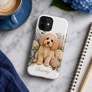 May include: A white smartphone case featuring a central illustration of a light brown curly-haired dog sitting amidst a cluster of blue, pink, and cream-colored hydrangea flowers. The name Honey is printed in a cursive brown script below the dog.