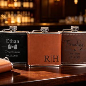 May include: Three rectangular metal flasks wrapped in black, tan, and brown leather-like material. The black flask features the name Ethan, a bowtie icon, and the date 06.26.2026. The tan flask displays the initials R|H. The brown flask shows the name Freddy, a diamond icon, and the date 02.14.2027.