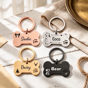 May include: Four bone-shaped metal pet ID tags with circular key rings. The top left is rose gold engraved with a bow, the name Sadie, and a paw print. The top right is silver with stars, a moon, the name Coco, and a paw print. The bottom left is gold with a deer, a tree, the name Daisy, and a paw print. The bottom right is black with a puppy, the name Bear, and a paw print.