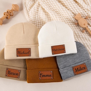 Peut inclure: Un ensemble de cinq bonnets en tricot dans les tons beige, blanc, ocre, marron et gris. Chaque bonnet comporte une étiquette rectangulaire en similicuir marron rivetée sur le revers, personnalisée avec les prénoms Michael, Matthew, Mason, Emma et Miles.
