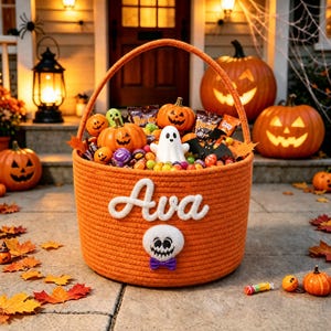 Peut inclure: Un panier rond en corde orange avec une anse, portant le prénom Ava brodé en blanc et un écusson en forme de crâne blanc avec un nœud papillon violet. Le panier est rempli de bonbons d'Halloween, de petites citrouilles, d'un fantôme et d'une chauve-souris.